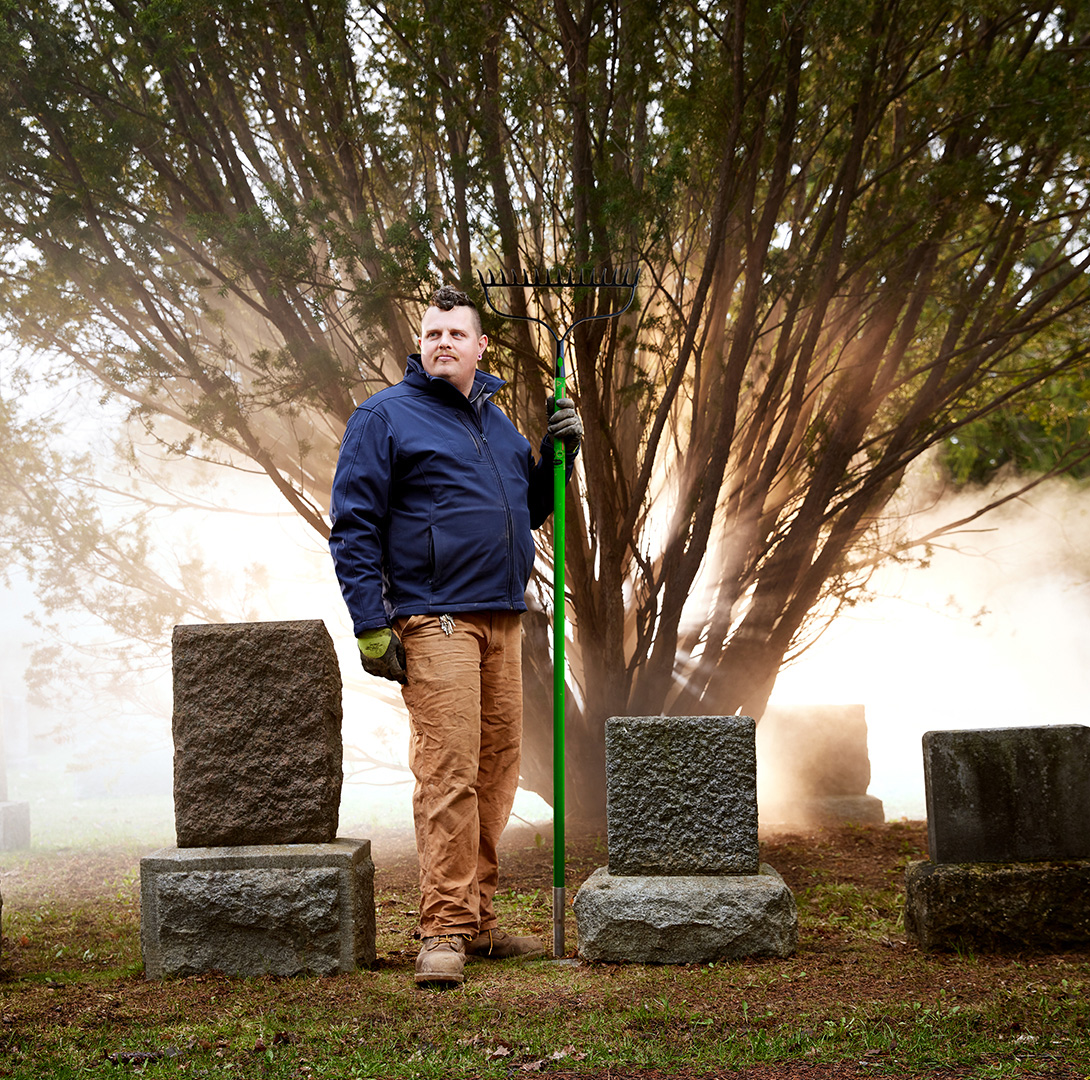 Cemetery Groundskeeper standing in front of a back-lit tree surrounded by tombstones.