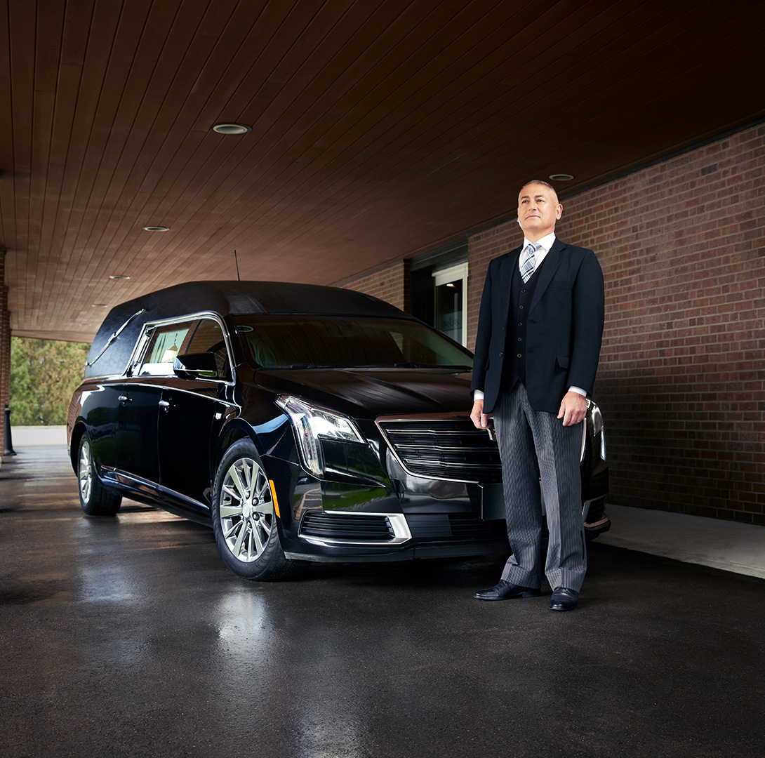 Assistant du directeur de funérailles debout devant un corbillard poli dans une allée.